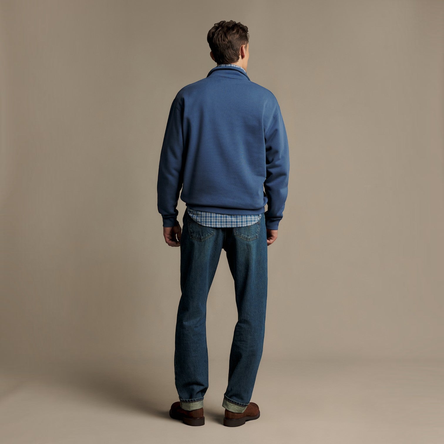 A man viewed from behind wearing a blue fleece pullover, a blue and white plaid shirt, dark blue jeans, and brown leather shoes, standing against a solid beige studio background.