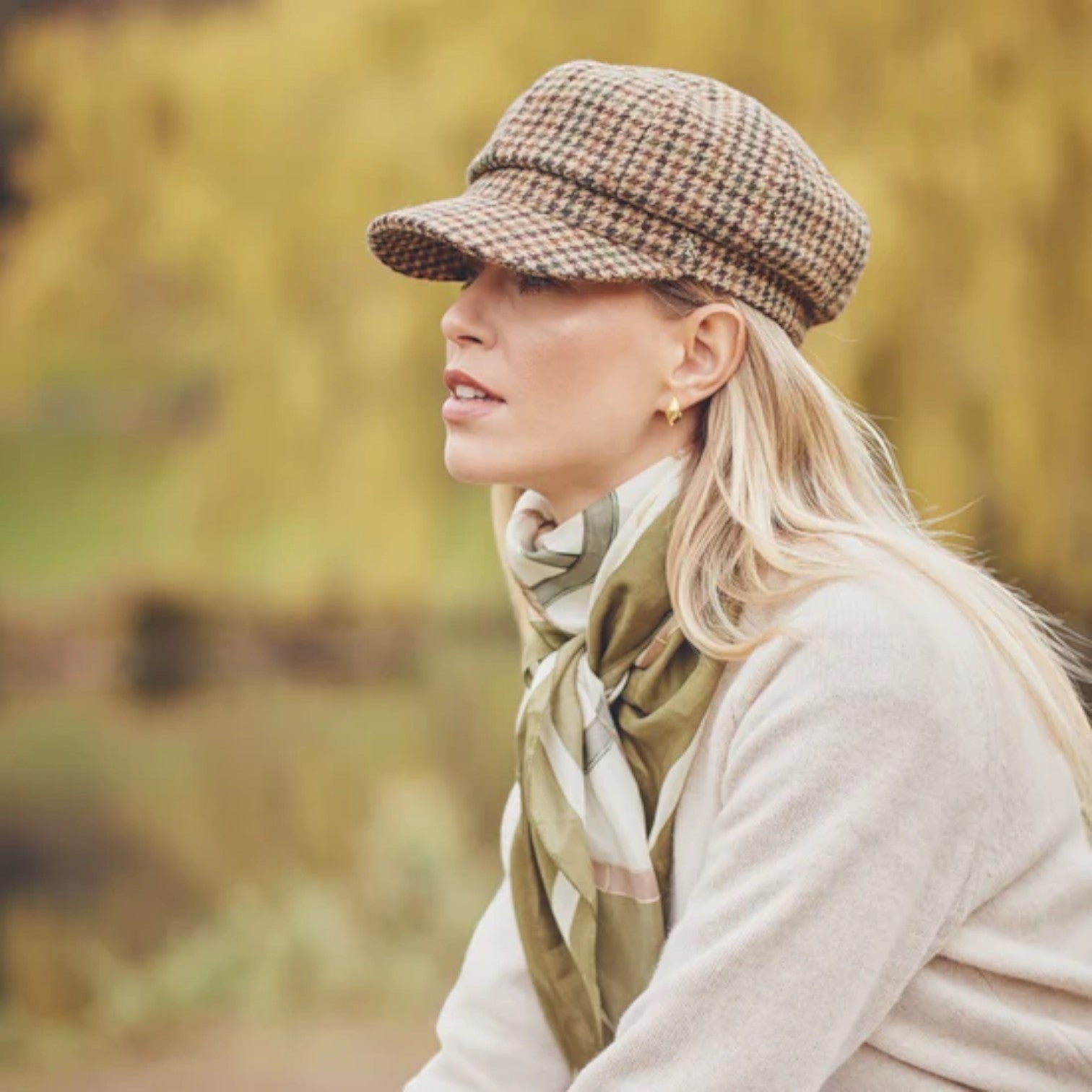 A woman in profile wearing a brown and beige houndstooth flat cap, a cream knit sweater, and an olive green scarf, set against a soft-focus outdoor background of yellow autumn foliage.