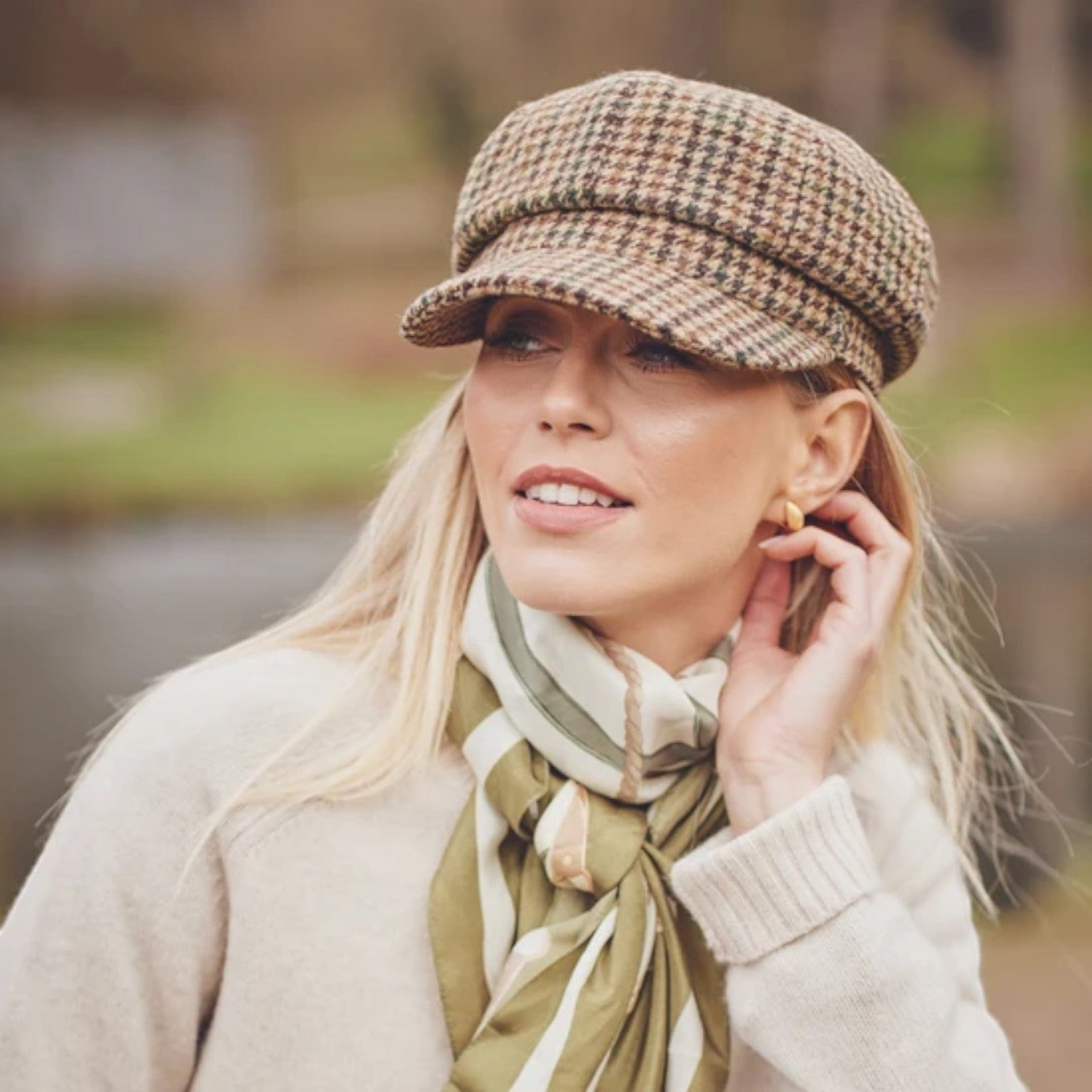 A woman wearing a brown and beige houndstooth newsboy cap, a cream knit sweater, and an olive green and cream striped scarf, posing outdoors with a blurred natural background.
