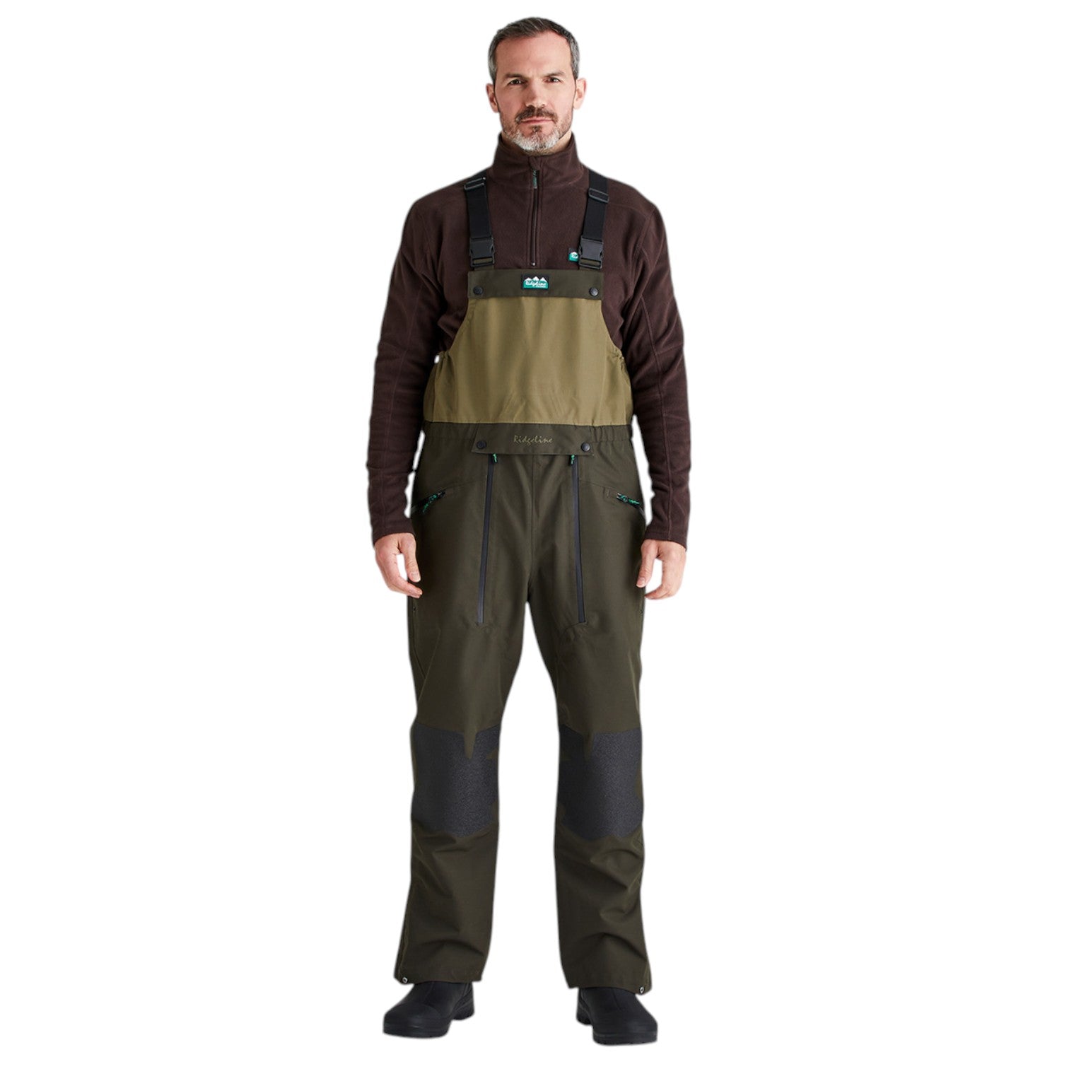 A man modeling a pair of outdoor bib overalls with a dark brown quarter-zip fleece underneath, shown against a plain white background.