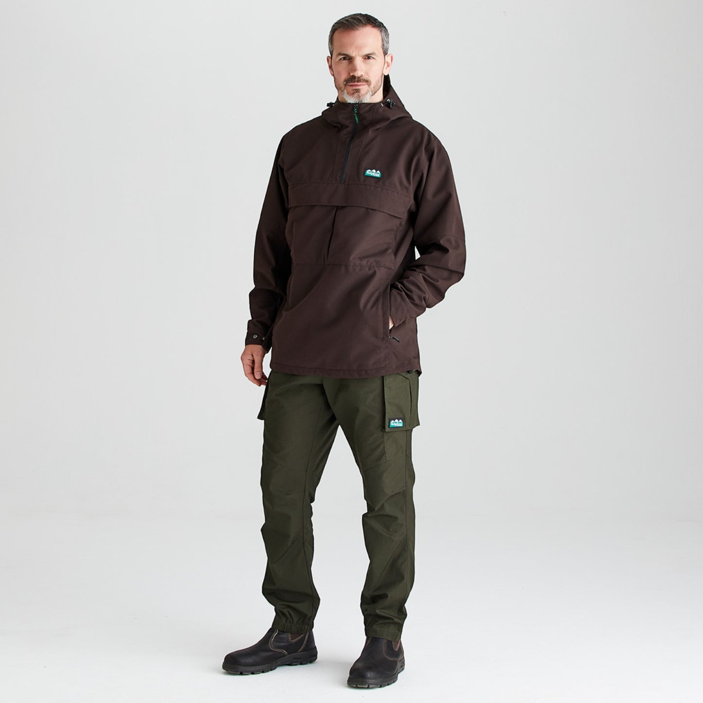 A man modeling a dark brown hooded pullover jacket with a small teal logo on the chest, paired with dark olive cargo pants and black boots, against a light gray studio background.