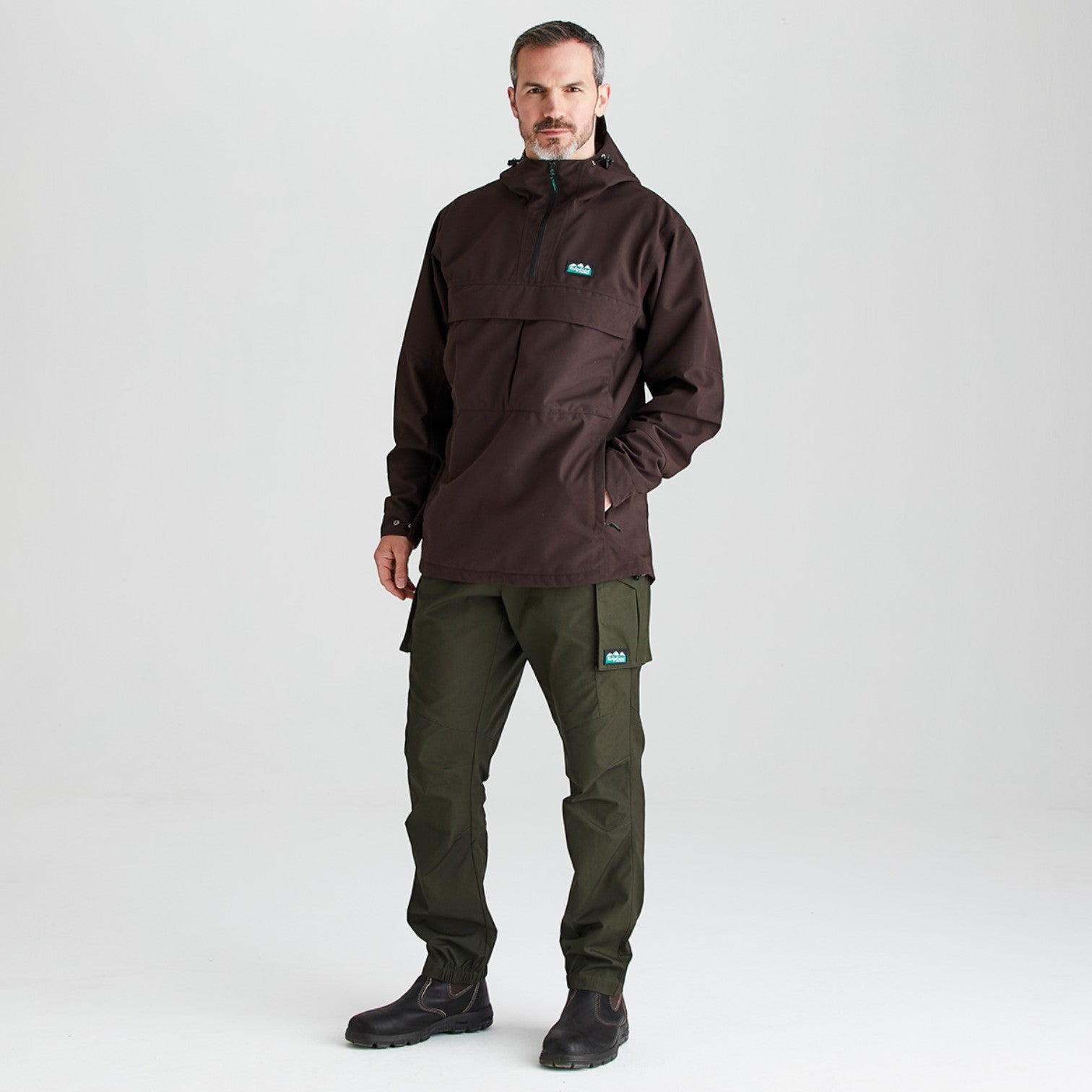 A man modeling a dark brown hooded pullover jacket with a small teal logo on the chest, paired with dark olive cargo pants and black boots, against a light gray studio background.