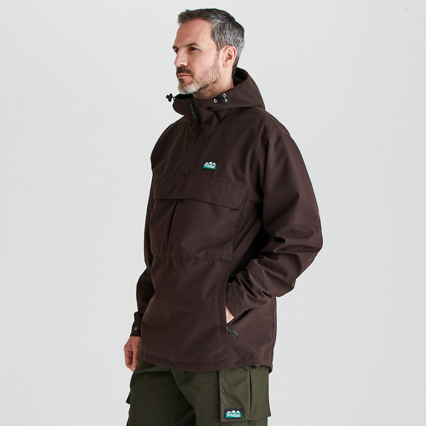 A man wearing a dark brown hooded anorak jacket with a small blue and white mountain logo on the left chest, paired with olive green pants, photographed against a light gray studio background.