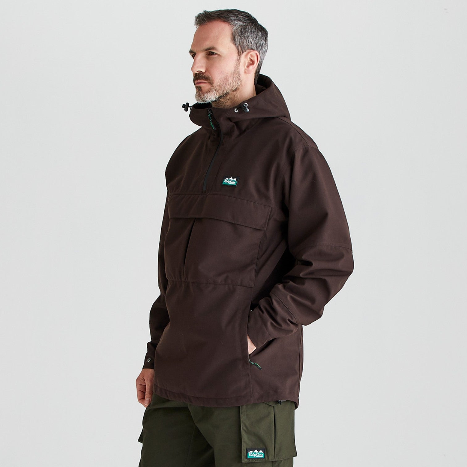 A man wearing a dark brown hooded anorak jacket with a small blue and white mountain logo on the left chest, paired with olive green pants, photographed against a light gray studio background.
