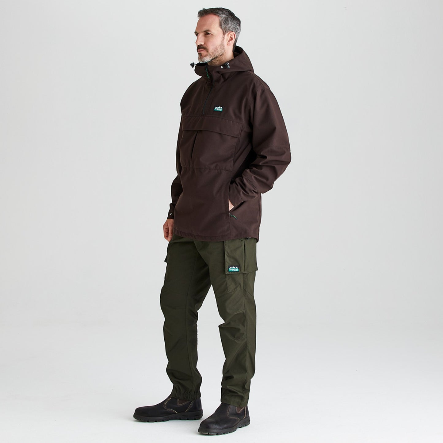 A man wearing a dark brown hooded jacket and dark olive green pants, standing in profile against a light gray studio background.