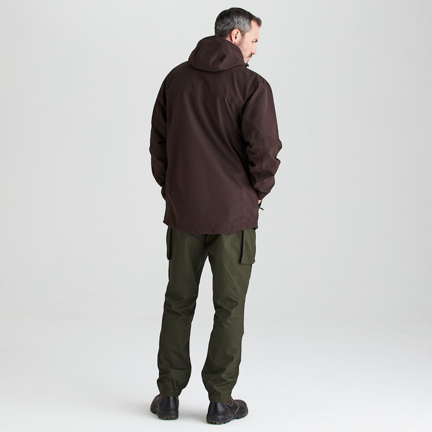 A man shown from the back wearing a dark brown hooded jacket and olive green cargo pants, standing against a light gray studio background.