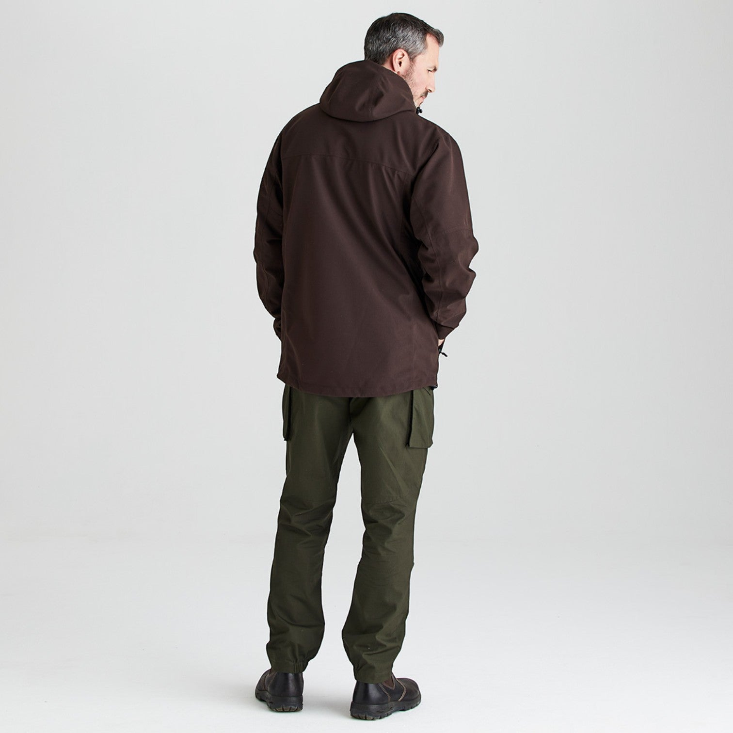 A man shown from the back wearing a dark brown hooded jacket and olive green cargo pants, standing against a light gray studio background.