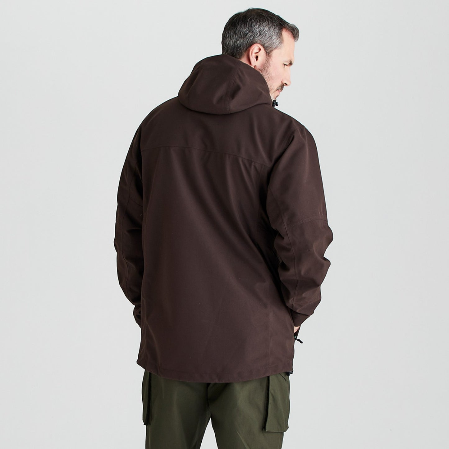 A man viewed from behind wearing a dark brown hooded jacket and olive green cargo pants, standing against a light gray studio background.