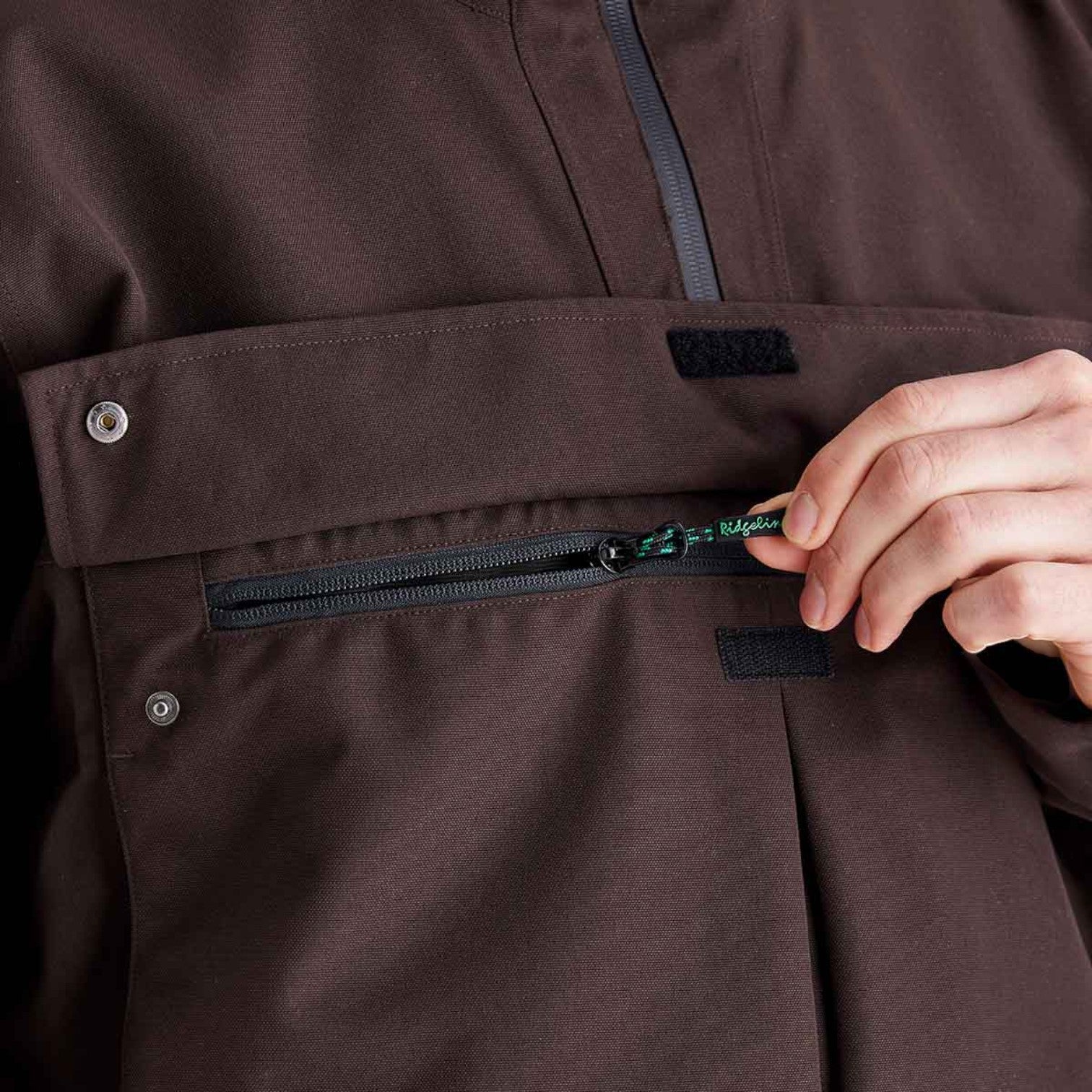 Close-up of a dark brown technical jacket with a hand unzipping a front flap pocket.