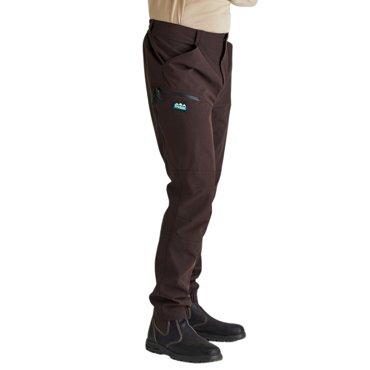 A side profile view of a person wearing dark brown technical pants with a zippered pocket on the thigh, paired with black boots and a beige shirt, against a plain white background.