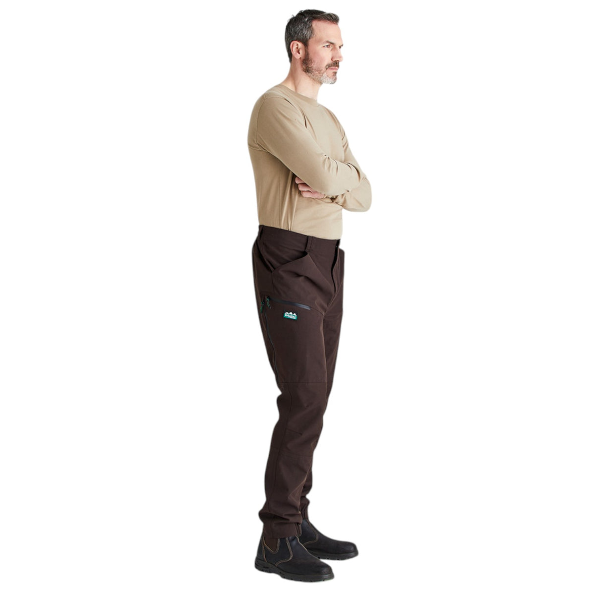A man in a tan long-sleeve shirt and dark brown pants stands in profile against a plain white background, with his arms crossed. The pants have a small light blue logo on the left thigh.