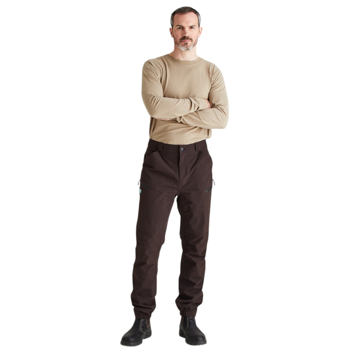 A man wearing a beige long-sleeve crewneck shirt and dark brown cargo pants with black boots, standing against a solid white background.