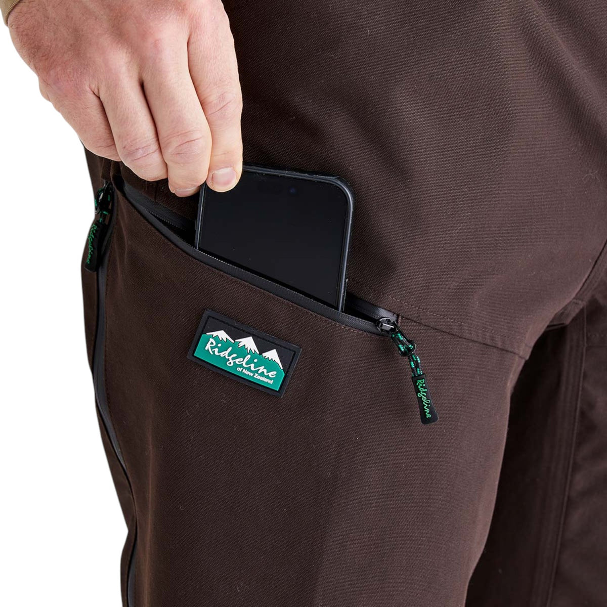 Close-up of a person's hand pulling a smartphone out of a zippered pocket on brown outdoor pants. A green and white Rideline logo patch is visible on the pocket.