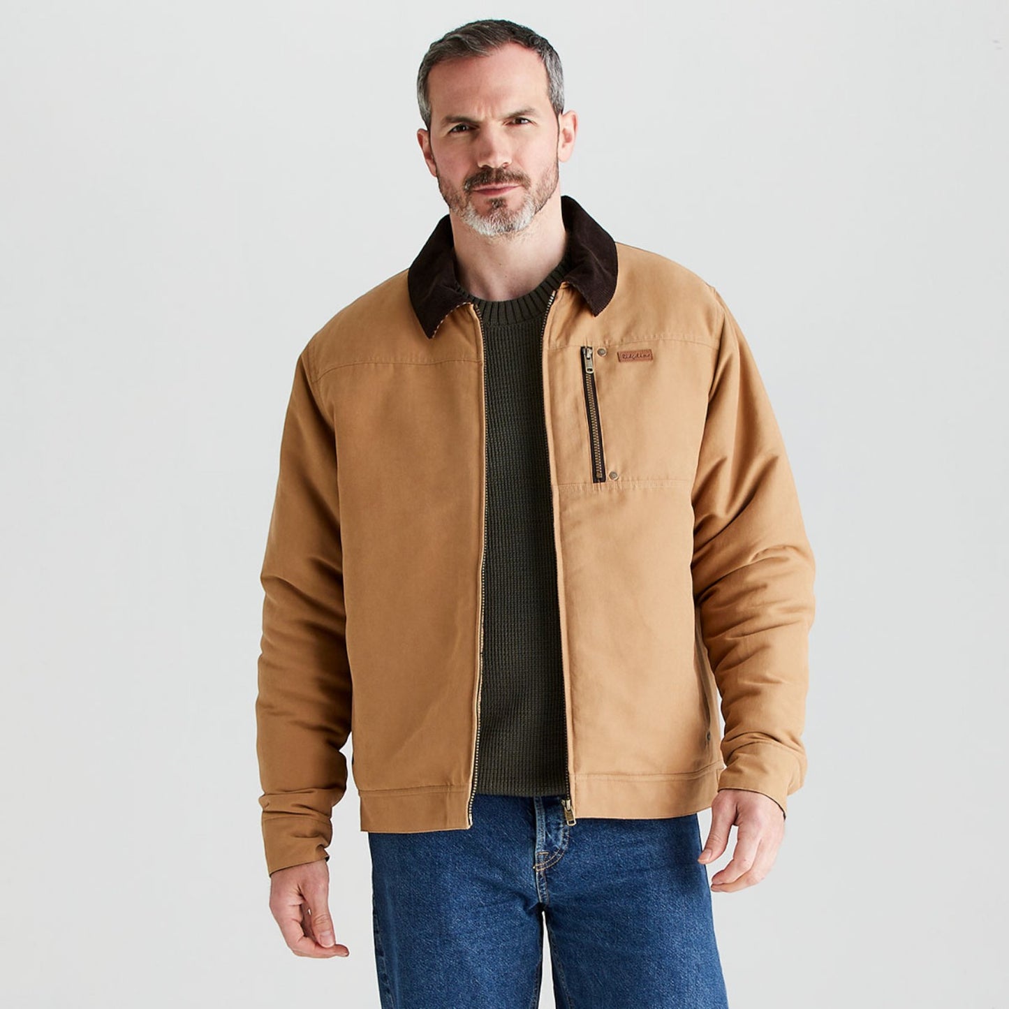 A man wearing a tan work jacket with a dark brown collar, layered over a dark green sweater and blue jeans, standing against a light gray studio background.