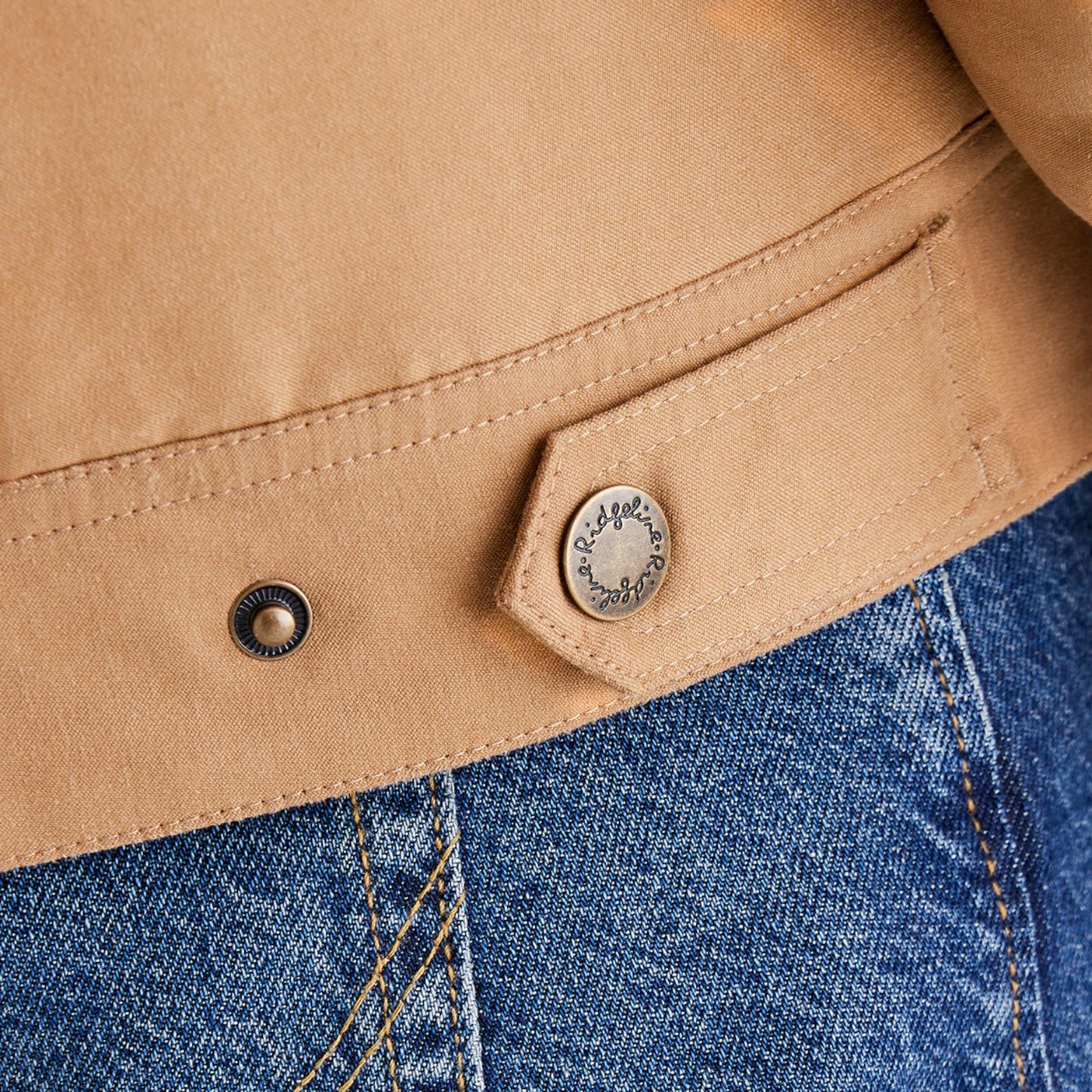 Close-up of a tan suede or nubuck jacket detail with a snap-button flap and a metal snap, shown next to blue denim.