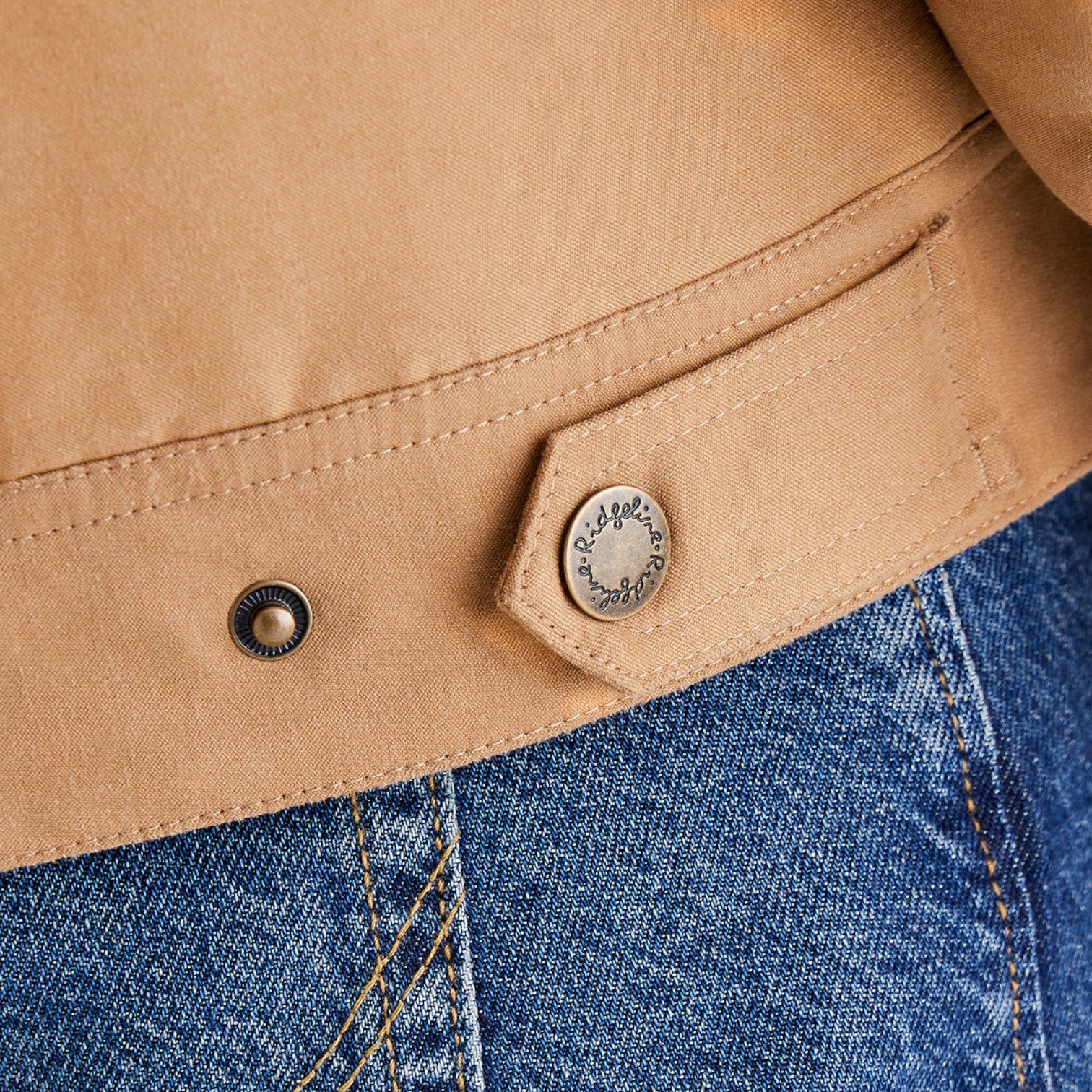Close-up of a tan suede or nubuck jacket detail with a snap-button flap and a metal snap, shown next to blue denim.