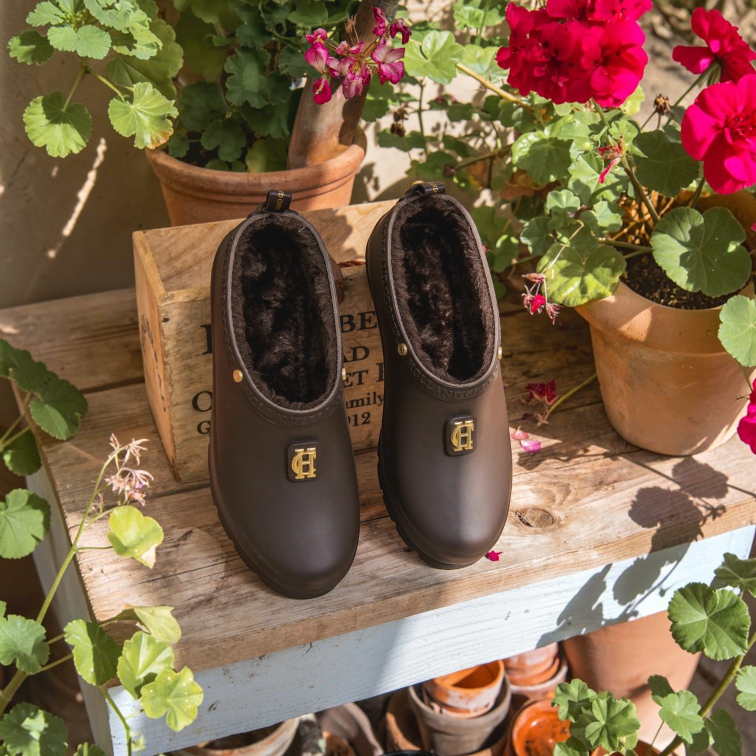 A pair of dark brown waterproof clogs with plush brown faux fur lining and a gold metal 'H' logo on the side, displayed on a rustic wooden crate surrounded by potted red flowers and green plants.