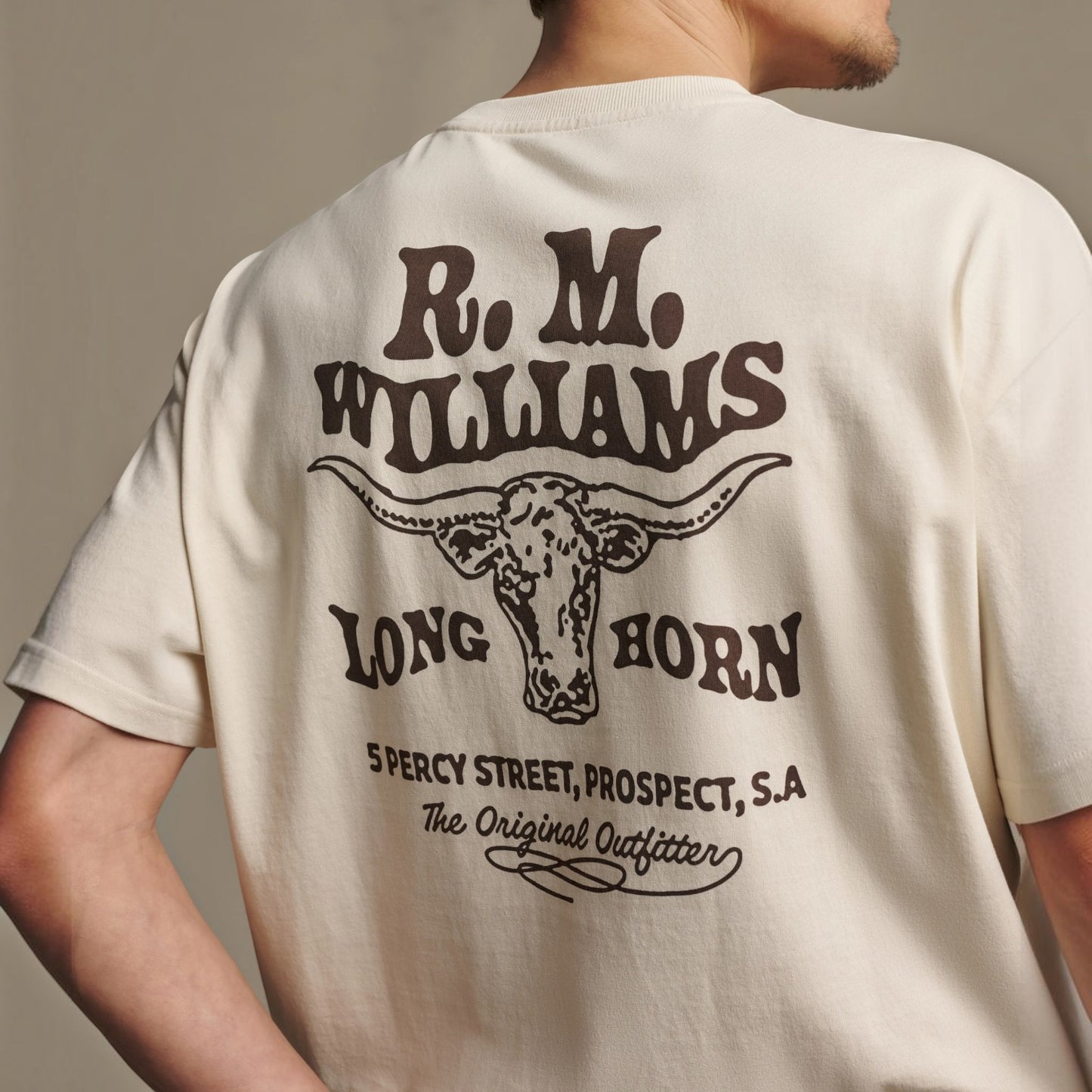 A man wearing a cream-colored t-shirt, viewed from the back. The shirt features a large dark brown graphic print of a longhorn steer's head with the text 'R.
