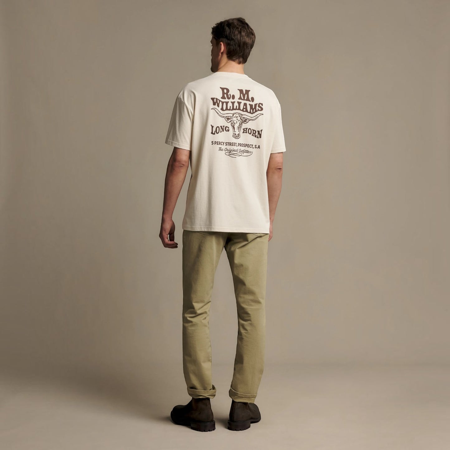 A man stands with his back to the camera wearing an off-white t-shirt with a dark brown graphic print on the back, paired with khaki pants and dark shoes, against a solid beige studio background.