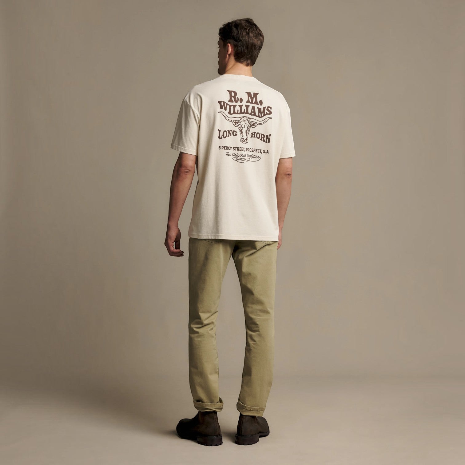 A man stands with his back to the camera wearing an off-white t-shirt with a dark brown graphic print on the back, paired with khaki pants and dark shoes, against a solid beige studio background.