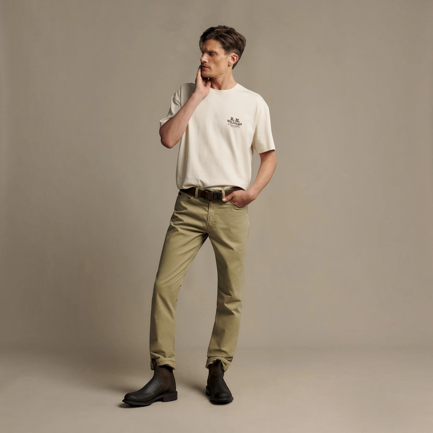 A man modeling a cream short-sleeve crewneck t-shirt with a small black chest logo, paired with olive green trousers and black boots, standing against a solid beige studio backdrop.