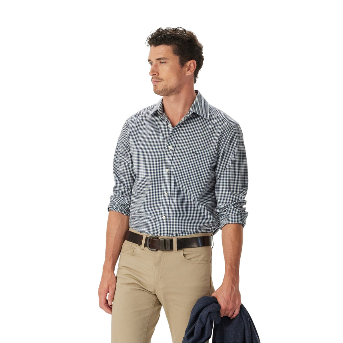 A man wearing a blue and white gingham long-sleeve button-down shirt, khaki pants, and a dark belt, holding a navy jacket against a solid white background.