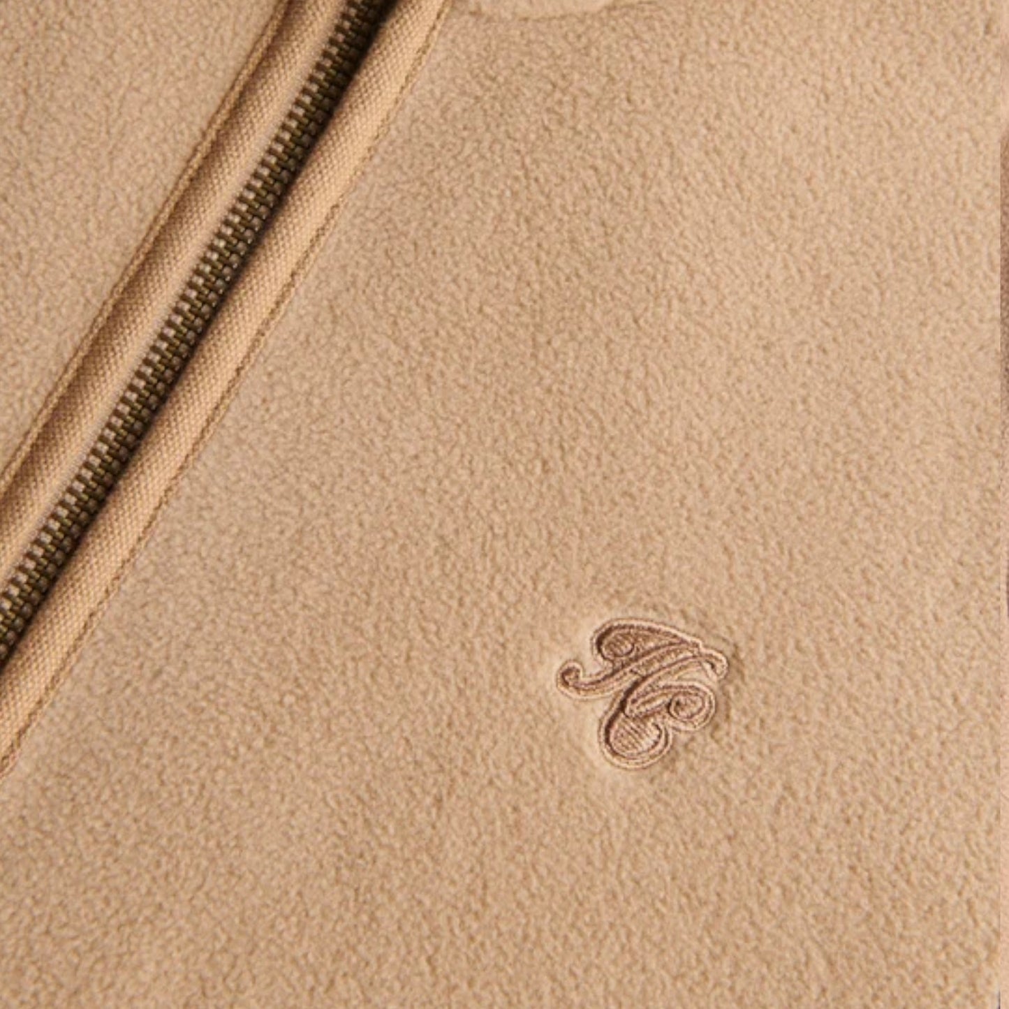 Close-up of a beige textured leather or suede surface with a diagonal zipper and a small tonal embroidered monogram logo.