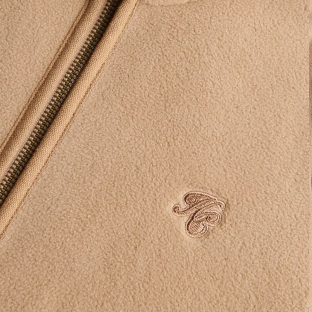 Close-up of a beige textured leather or suede surface with a diagonal zipper and a small tonal embroidered monogram logo.