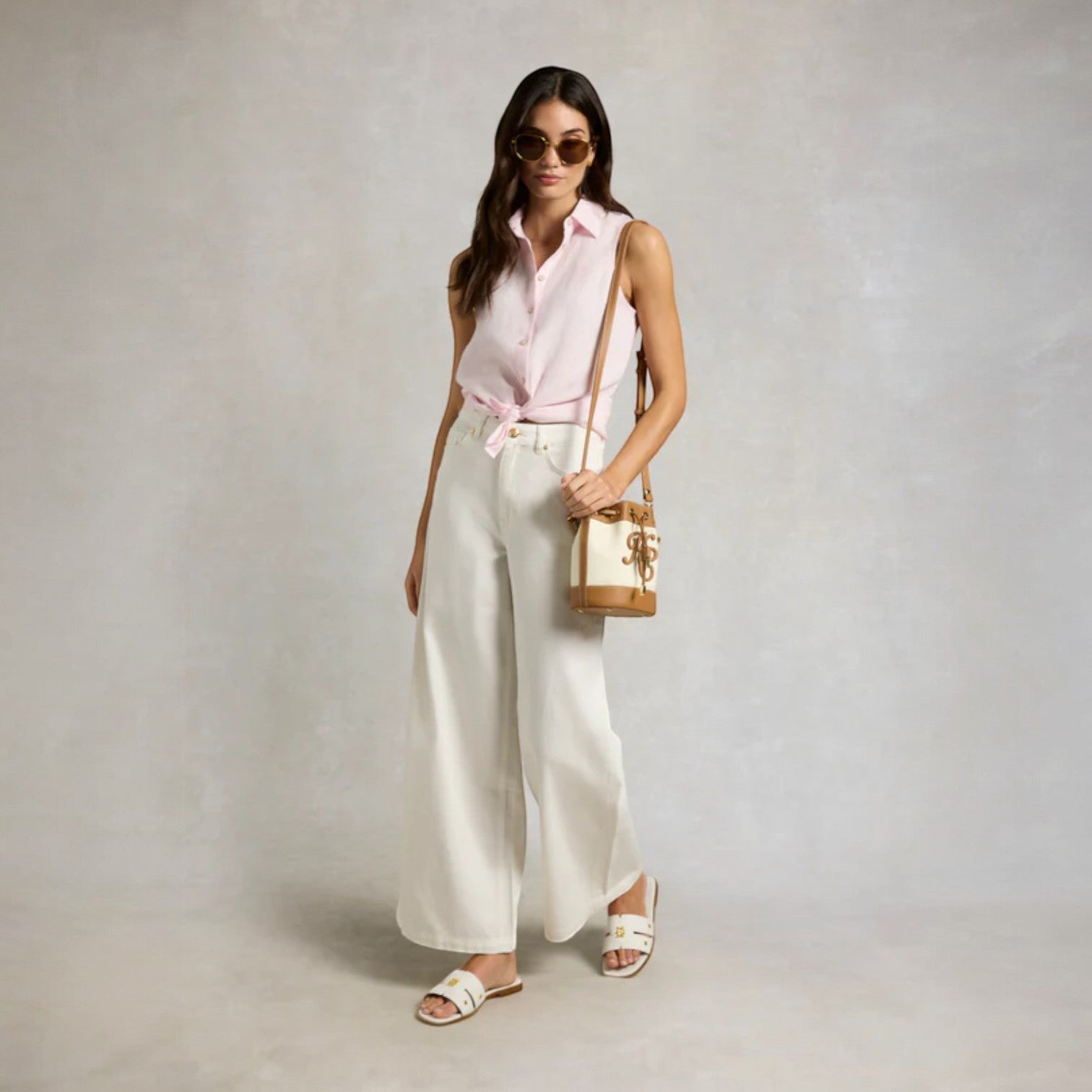 Woman wearing a sleeveless pink blouse and white pants, holding a handbag against a plain background