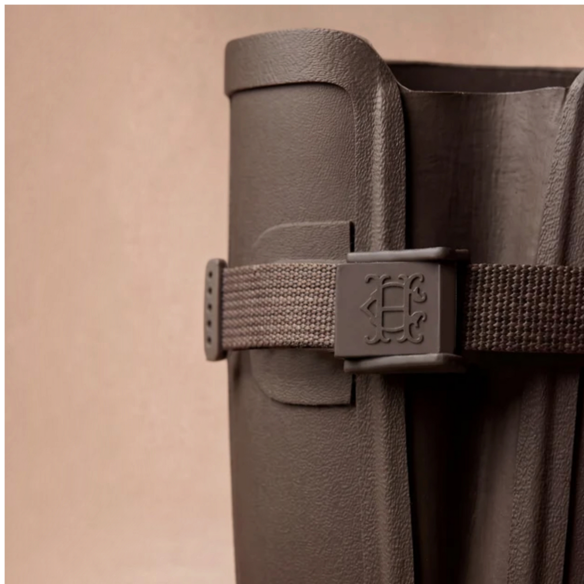 Close-up of a dark brown textured leather boot with a matching woven strap and embossed logo buckle, set against a solid beige background.