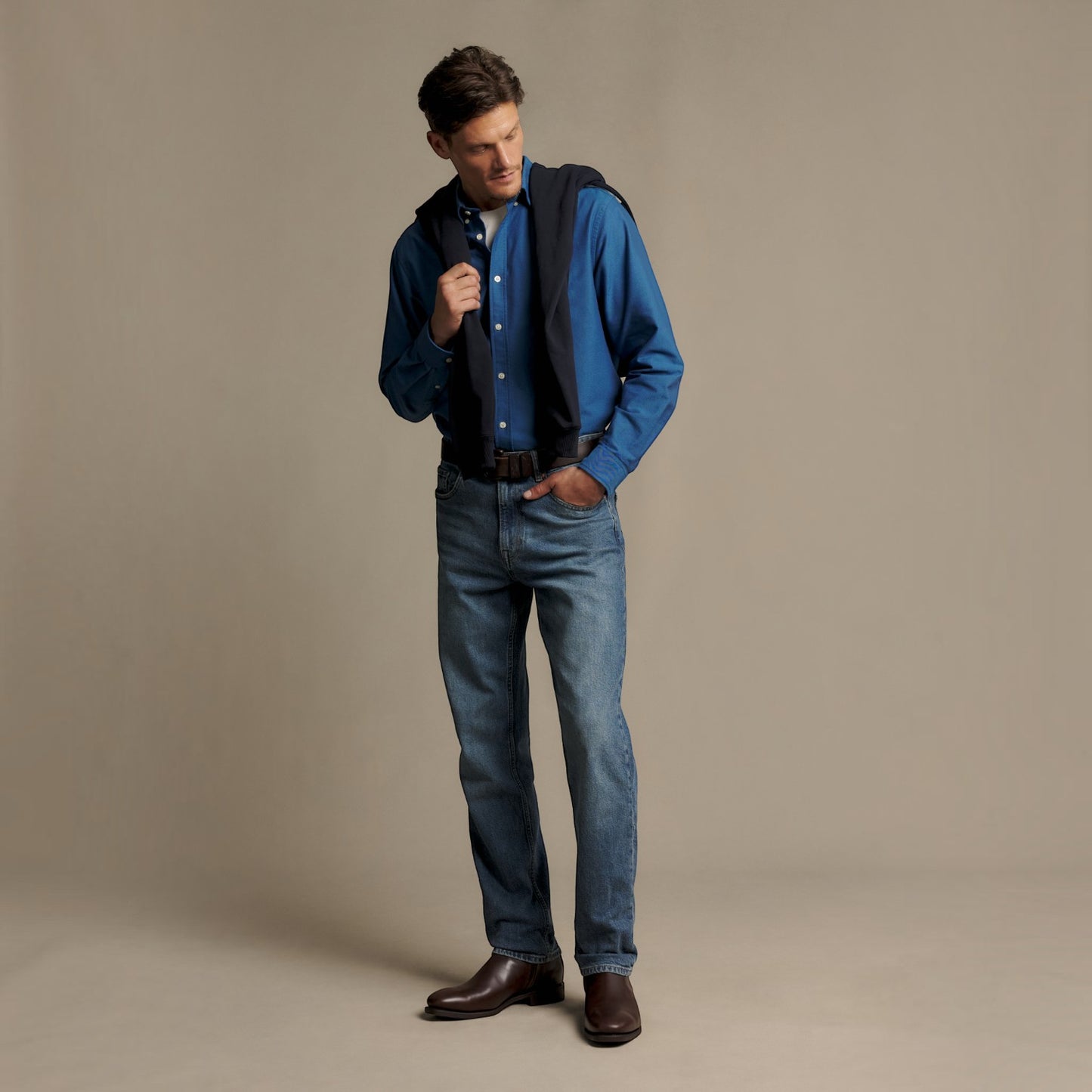 A man modeling a blue long-sleeve button-down shirt, blue jeans, and brown leather shoes, with a dark vest draped over his shoulders, against a solid beige studio background.