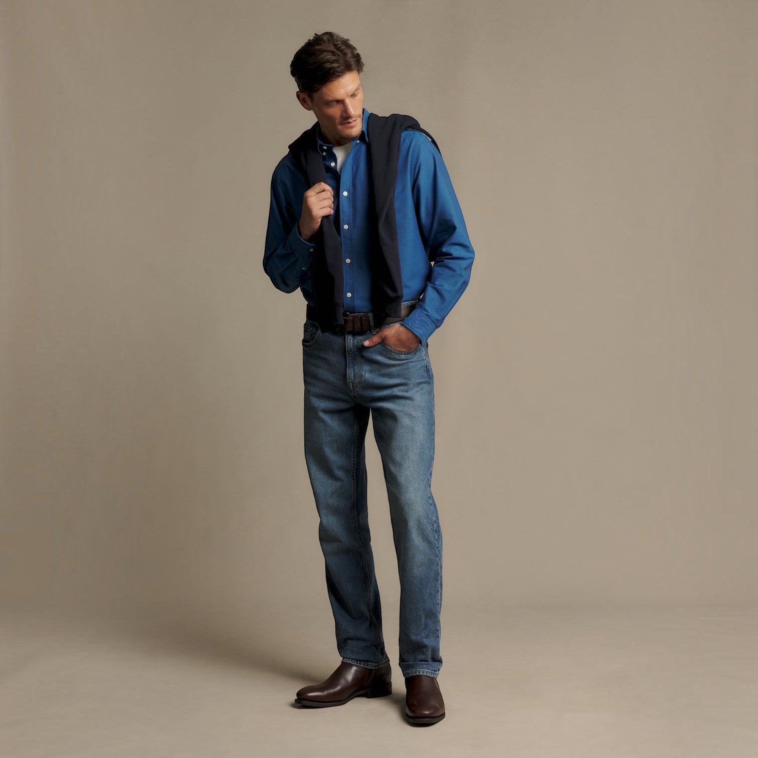 A man modeling a blue long-sleeve button-down shirt, blue jeans, and brown leather shoes, with a dark vest draped over his shoulders, against a solid beige studio background.