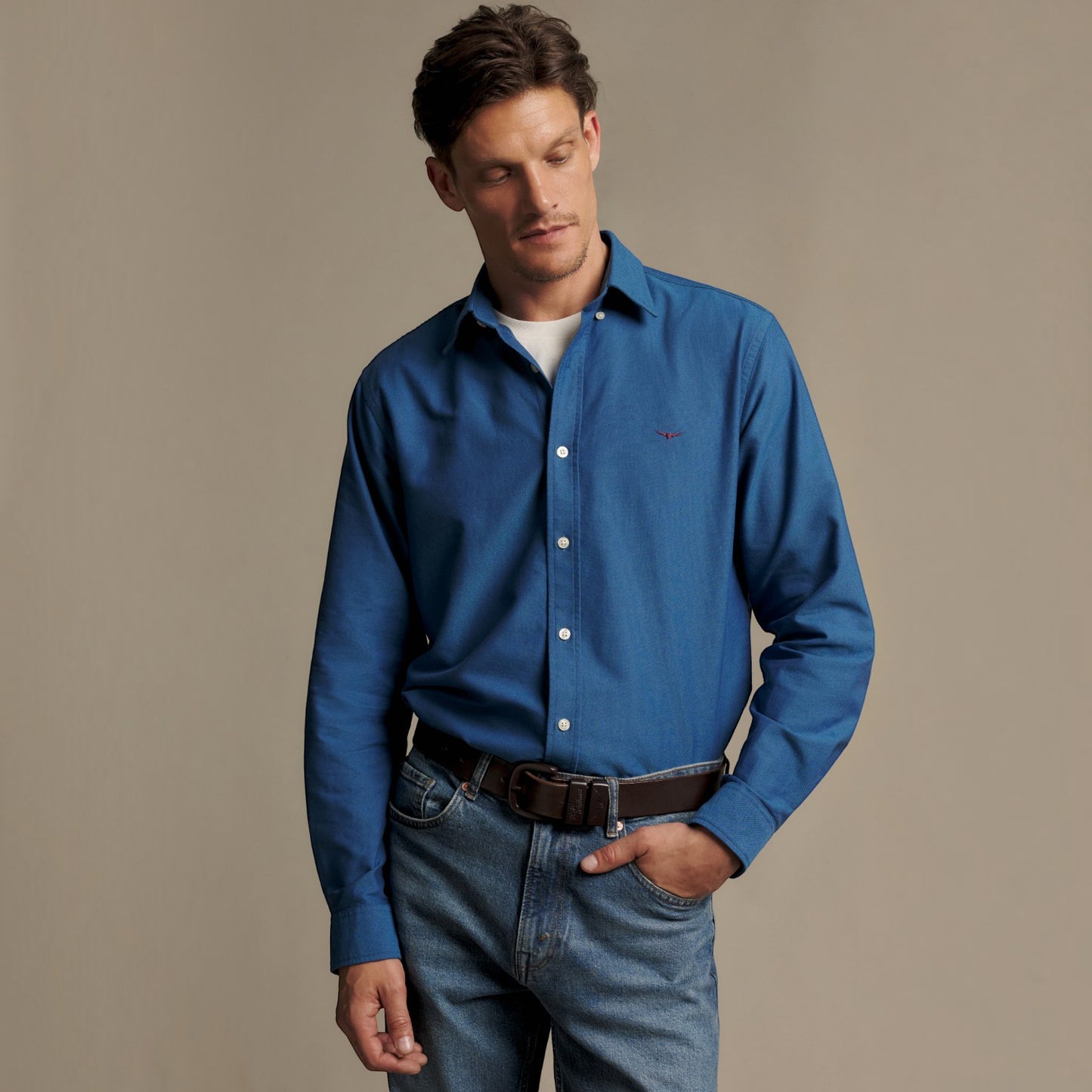 A man wearing a blue long-sleeve button-down shirt, blue jeans, and a brown belt, standing against a plain beige studio background.