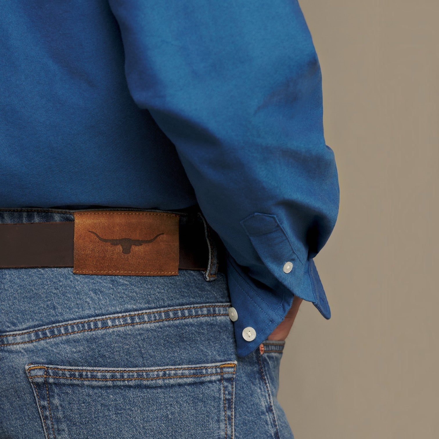 Close-up of a person wearing a blue long-sleeve button-up shirt tucked into blue jeans with a brown leather belt featuring a longhorn emblem.