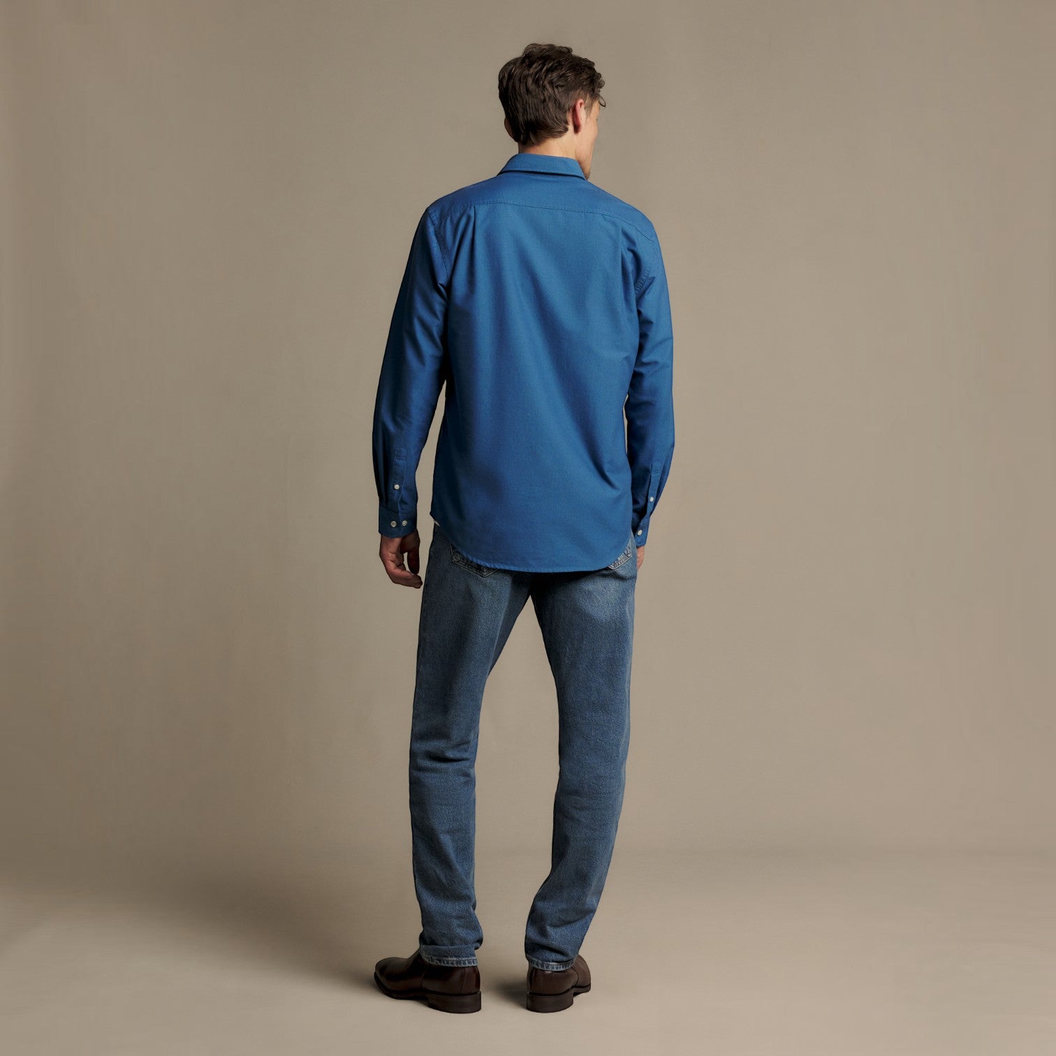 A man viewed from behind wearing a blue long-sleeve button-up shirt, blue jeans, and brown shoes, standing against a plain beige studio background.