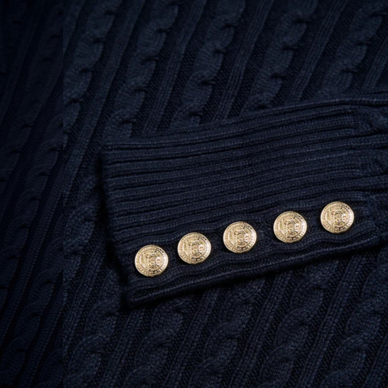 Close-up of a dark navy cable-knit garment with a ribbed cuff detail and five decorative gold buttons.
