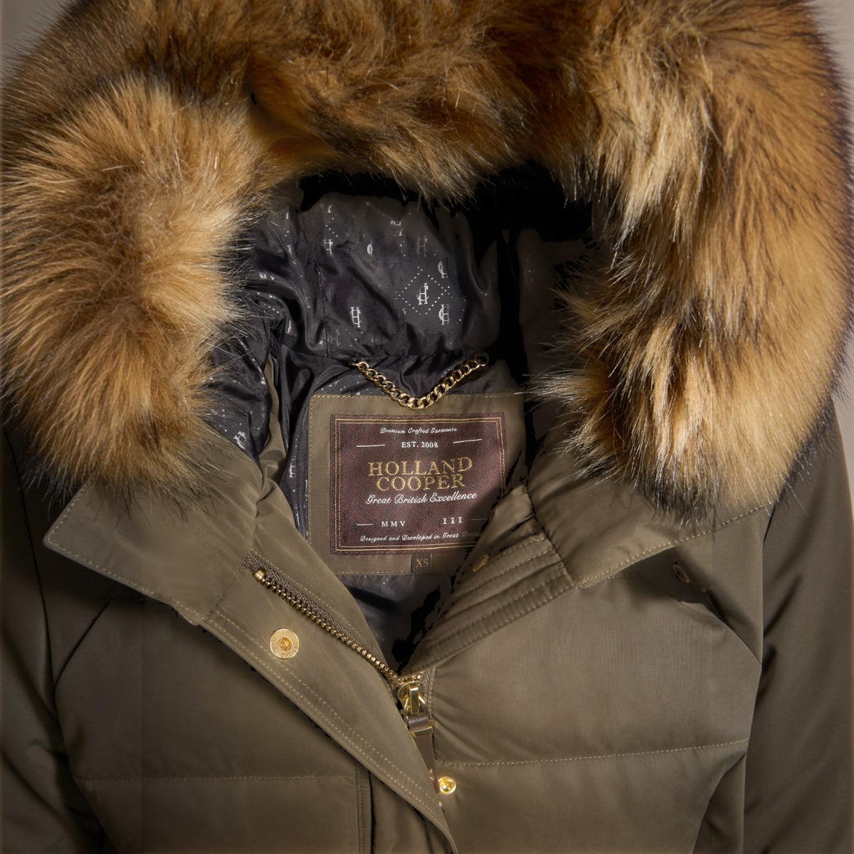 Close-up of a dark olive green puffer parka with a large brown and beige faux-fur hood trim, showing the interior lining and a Holland Cooper brand label.