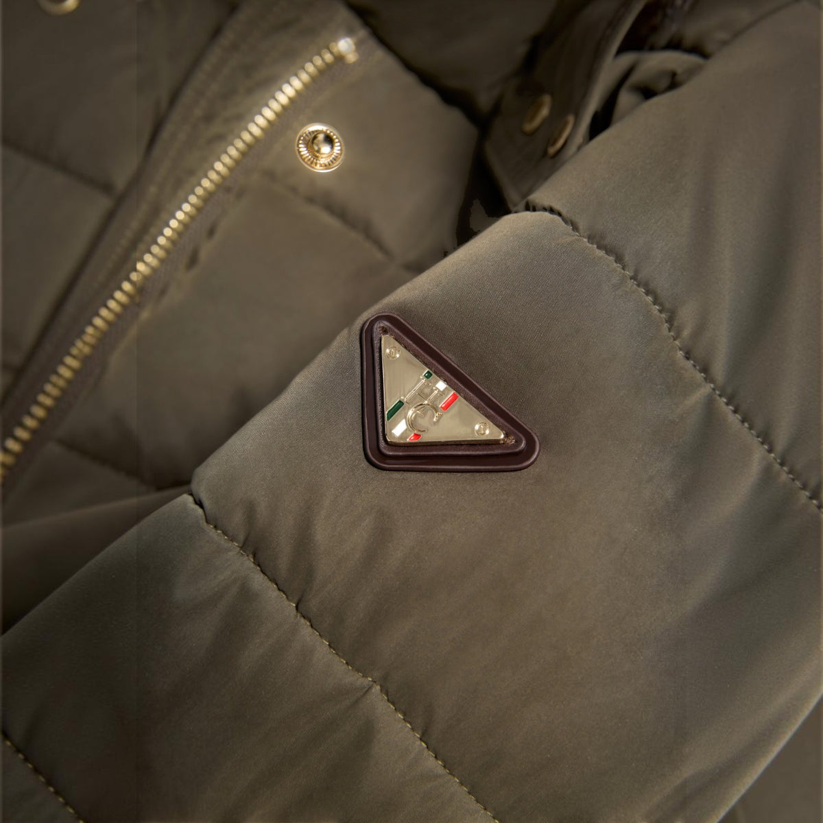 Close-up of a dark olive green puffer jacket showing the iconic triangular logo patch on the sleeve, alongside a gold zipper.