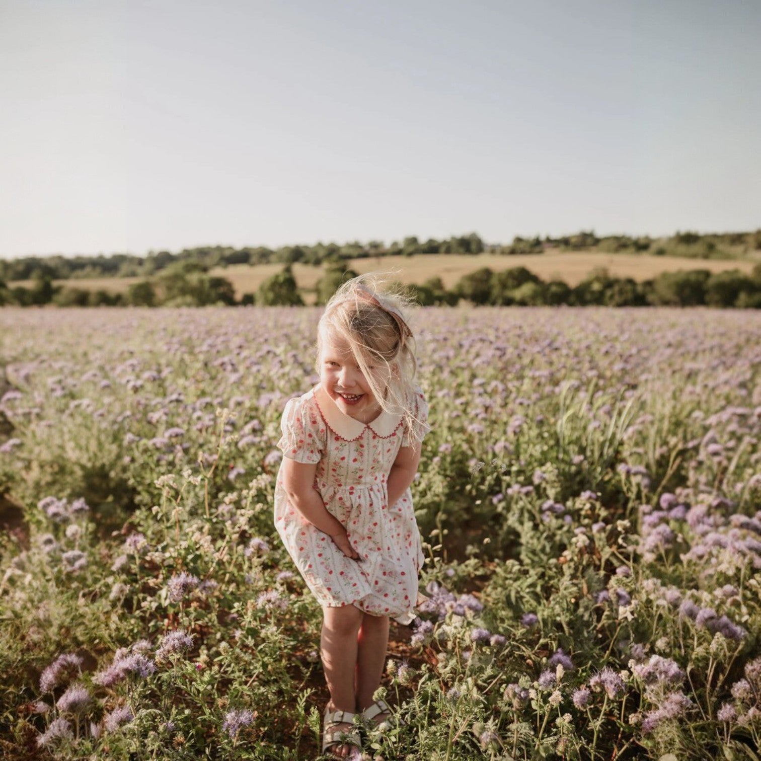 Young girl in a floral dress standing in a field of purple flowers with a clear sky.