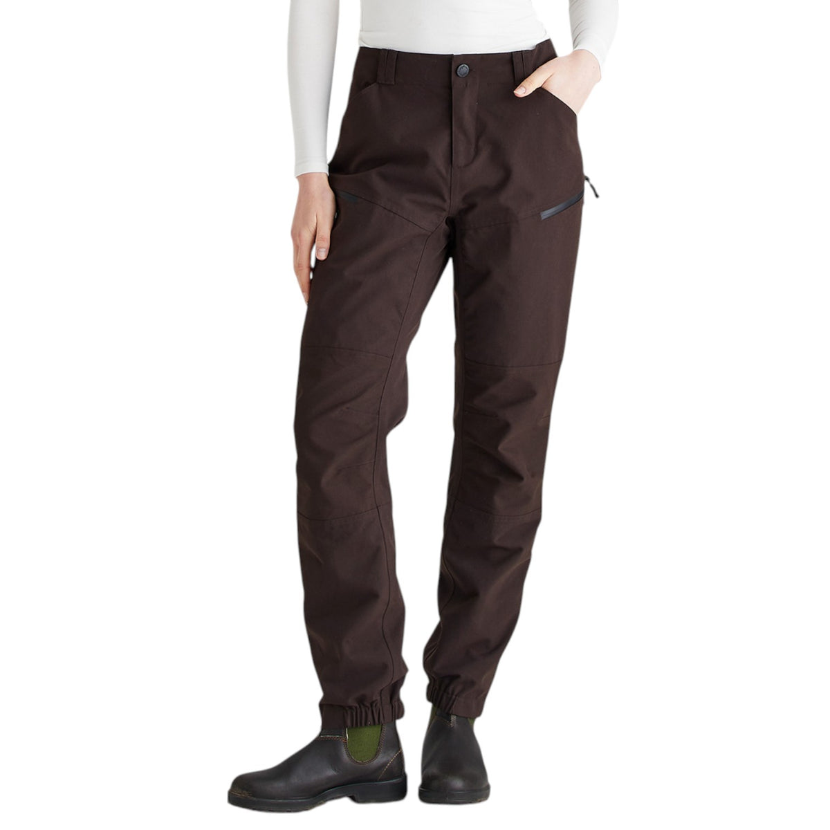 A person wearing dark brown technical pants with a zippered pocket and elastic cuffs, paired with black boots and a white long-sleeve top, shown against a plain white background.