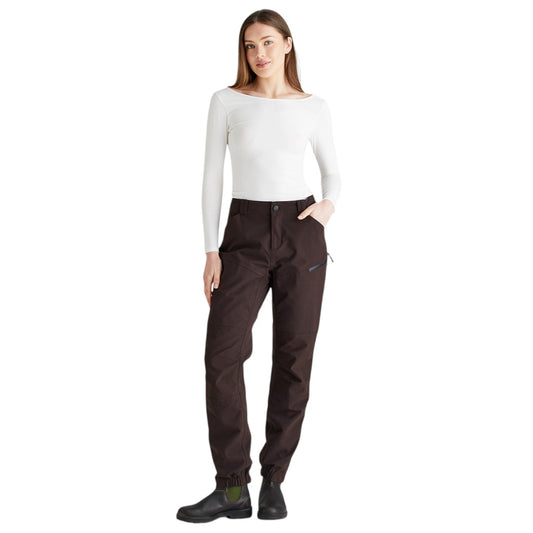 A woman modeling dark brown waterproof pants with a white long-sleeve top and black boots, standing against a solid white background.