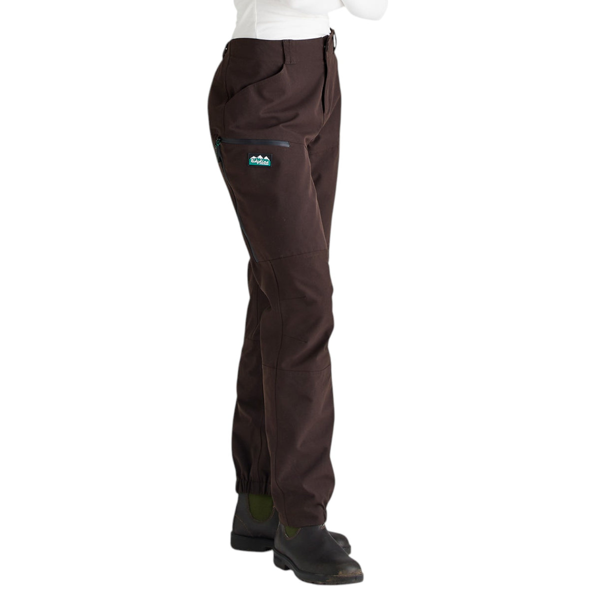 A side profile view of a person wearing dark brown technical pants with a small green and blue logo patch on the upper left thigh, paired with a white top and black boots, against a plain white background.