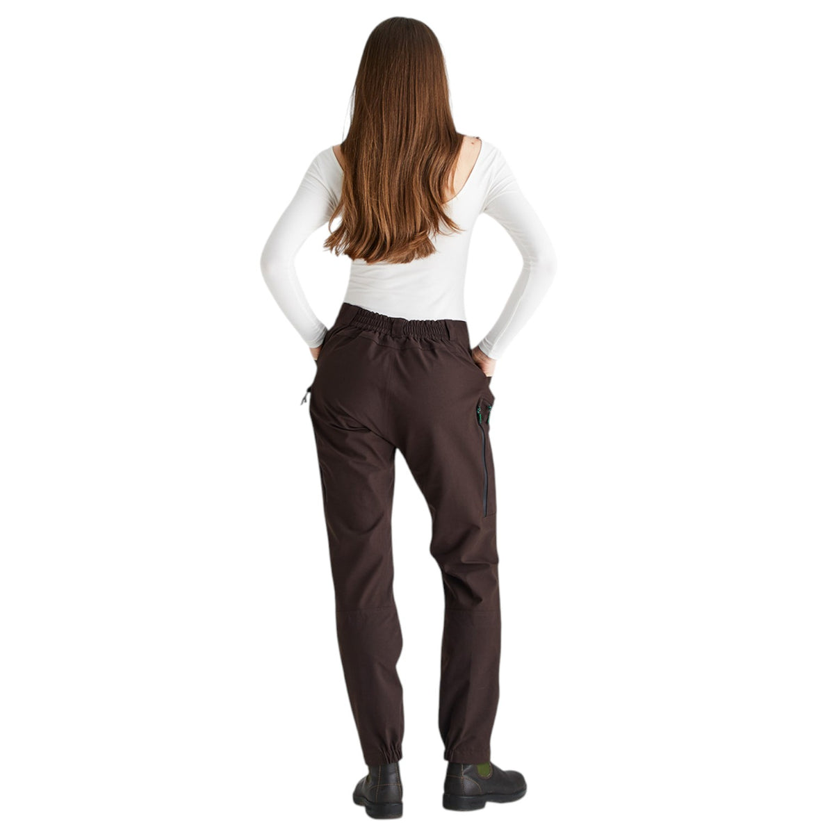 A woman viewed from the back wearing dark brown pants, a white long-sleeve top, and black shoes against a plain white background.