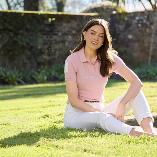 Woman sitting on grass wearing a pink polo shirt with text and white pants.