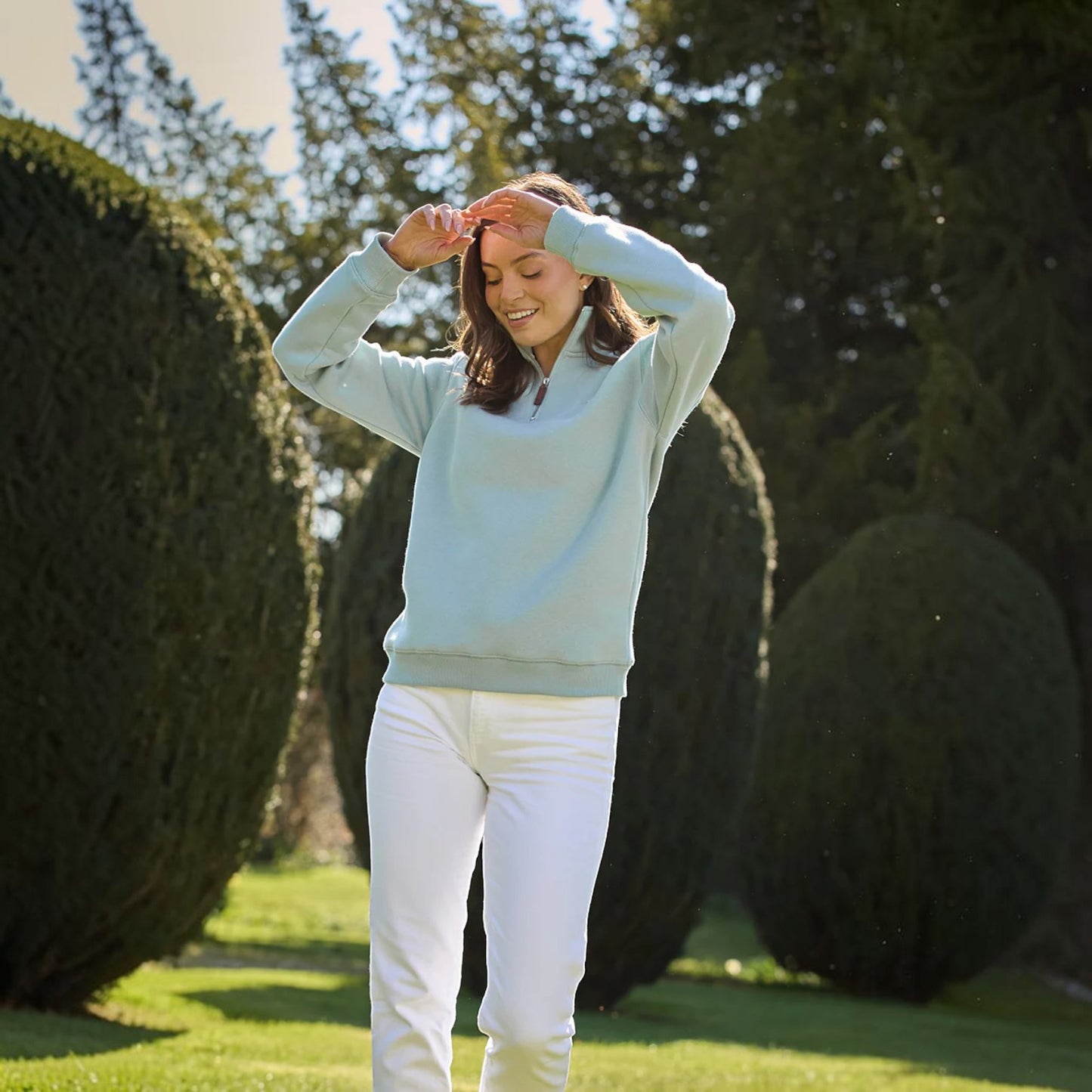 Woman in light blue sweater and white pants standing on a grassy area with trees in the background