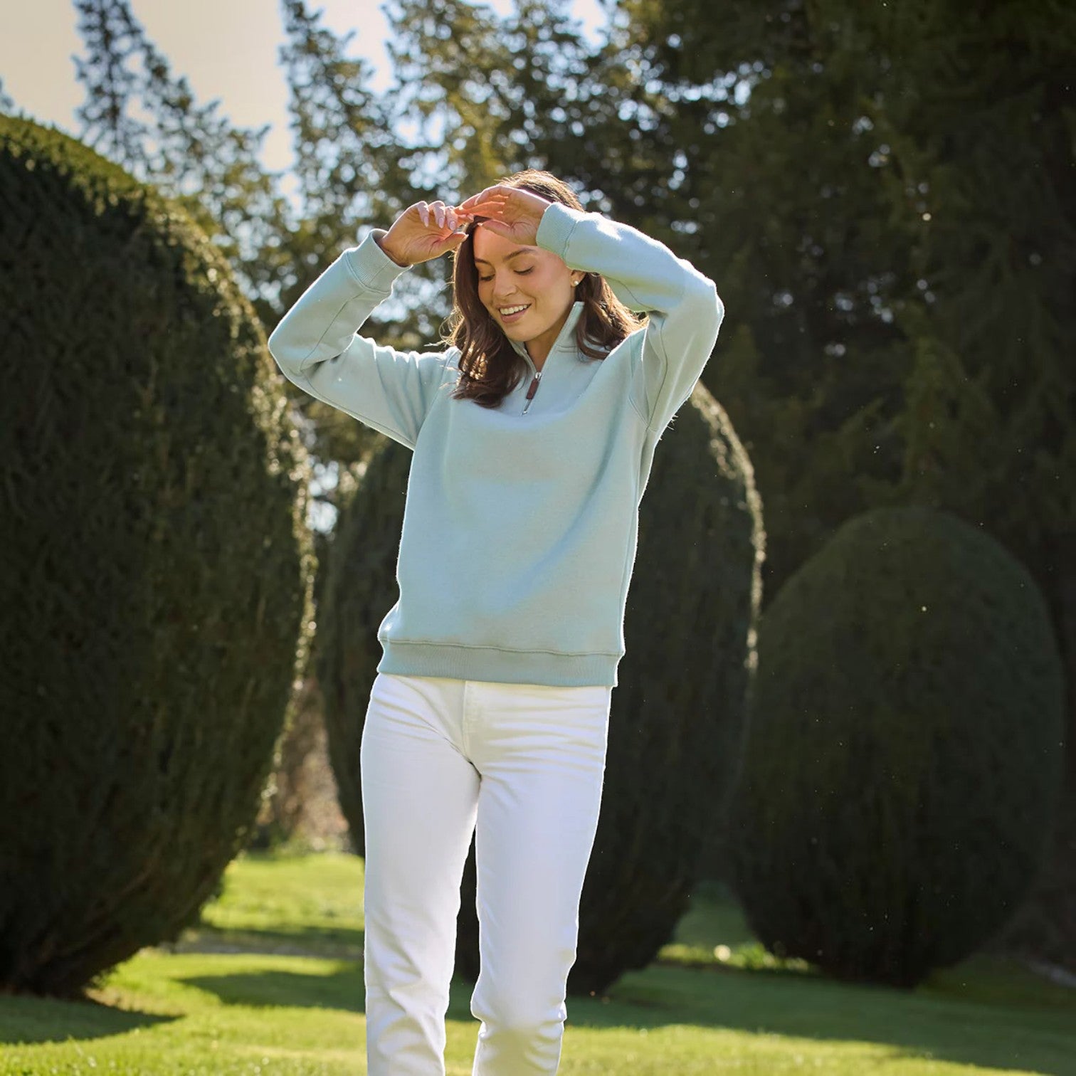Woman in light blue sweater and white pants standing on a grassy area with trees in the background