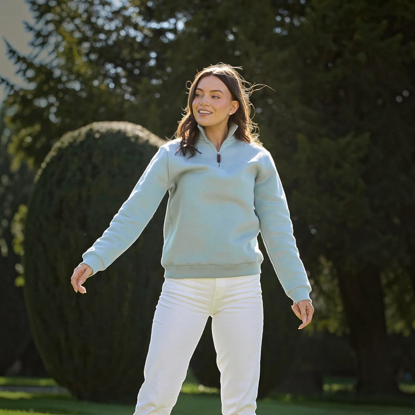 Woman wearing a light blue sweater and white pants standing outdoors with trees in the background