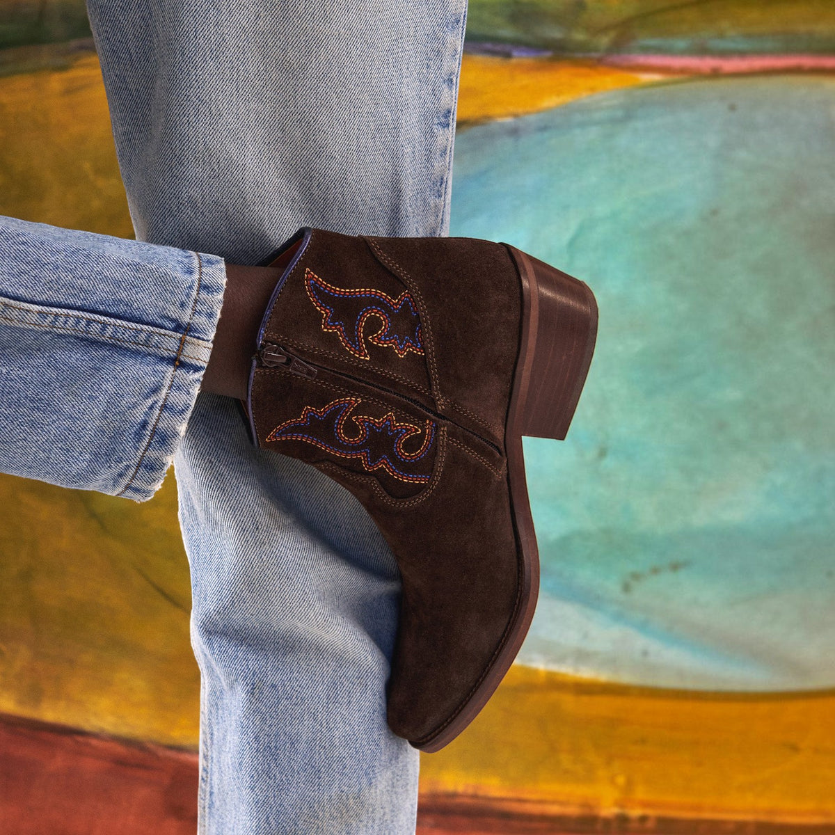 A person wearing light blue jeans and a brown suede western ankle boot with embroidered details, posed against an abstract painted background.