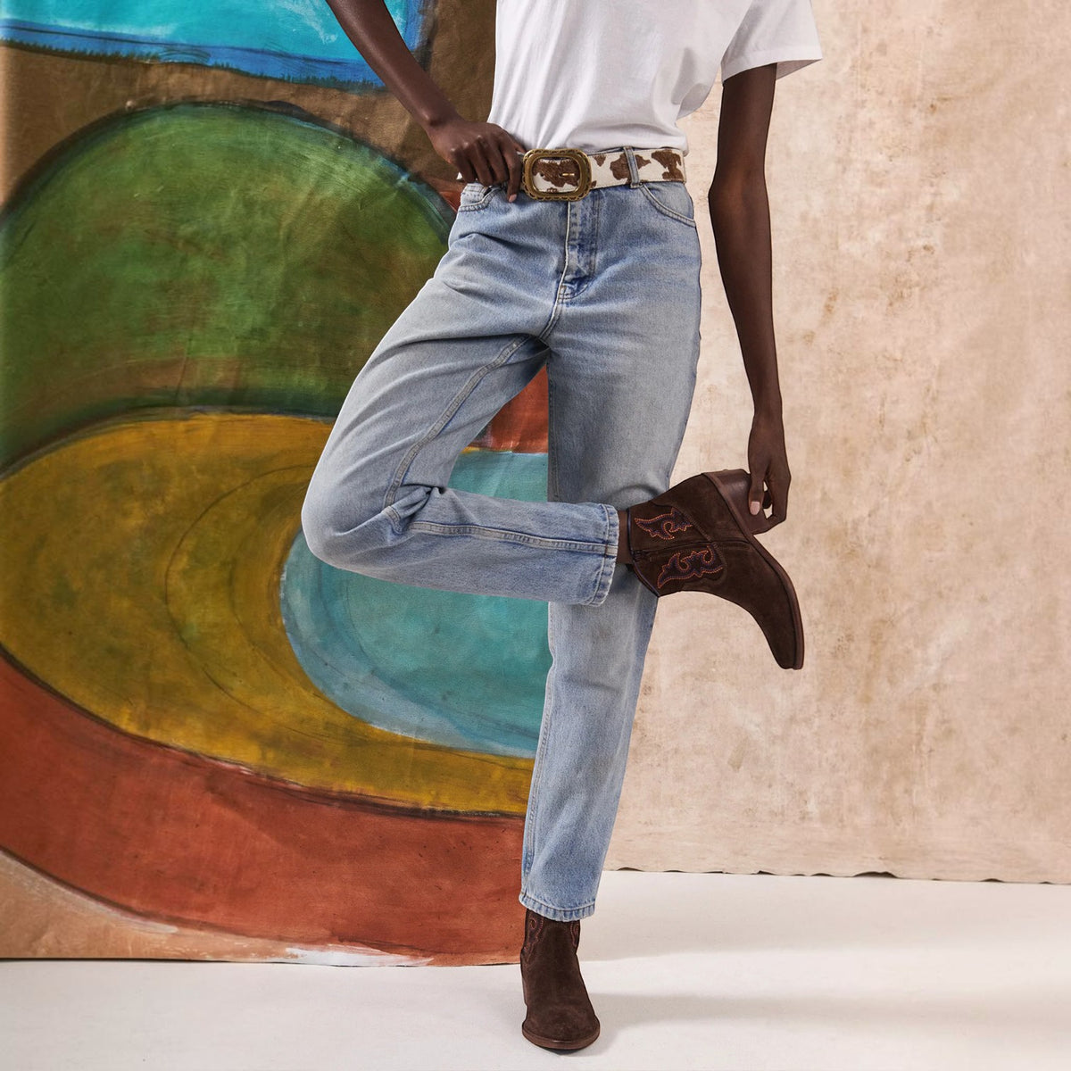 A model wearing light-wash blue jeans with a leopard-print belt and brown western boots, standing in front of a textured beige wall and a colorful abstract painting.