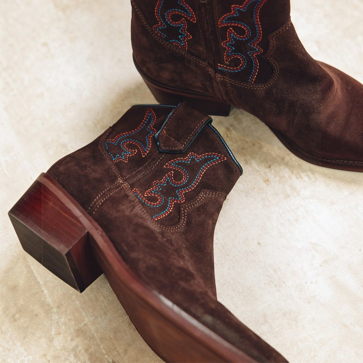 A pair of dark brown suede western boots with embroidered blue and red detailing, shown on a light beige stone surface.